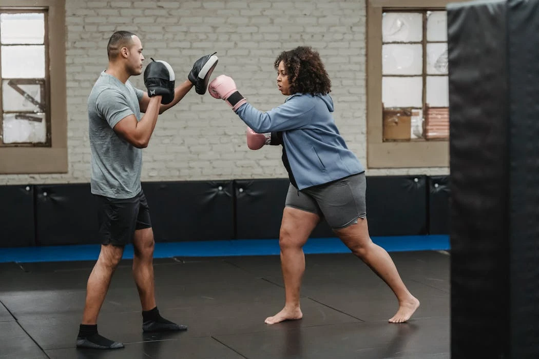 Side view of concentrated overweight African American female in boxing gloves hitting personal trainer during workout